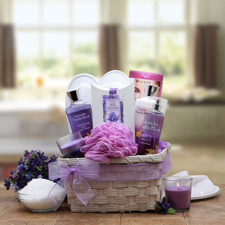 Lavender spa gift basket with bath and body products in a white wicker tray, including lotion, bath gel, body spray, bath salts, loofah, candle, slippers, and eye pads.